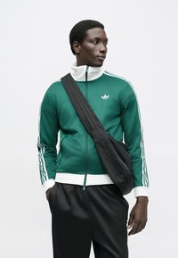Green zip-up sweatshirt with white accents and three stripes on sleeves, featuring a high collar and logo; worn with black pants and a black bag.
