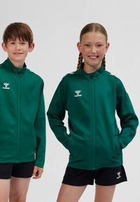 Gröna zip-up jackor gjorda av slätt material med vita axeldetaljer. Två barn står sida vid sida, båda klädda i svarta shorts.