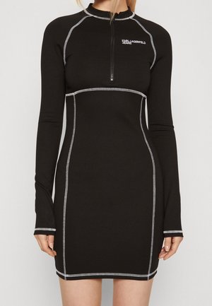 Černé dlouhé šaty s dlouhým rukávem z elastické látky, s zipem u krku, kontrastním bílým prošíváním podél švů a přiléhavým střihem.