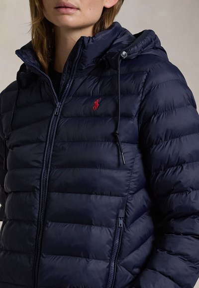 Naine kannab tumesinist tikitud sulejope kapuutsi ja punase tikitud hobuse logoga rinnal.