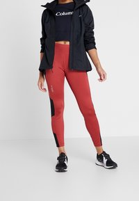 Sportoutfit bestaande uit een zwarte lichtgewicht jas, een marineblauwe crop top en strakke rode leggings met zwarte zijkanten en logo-accents. Zwarte sneakers.