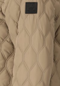 Gewatteerde beige stof met diamantvormige stiksels en een zwart label met reliëf van een berglogo en de tekst "Whistler".