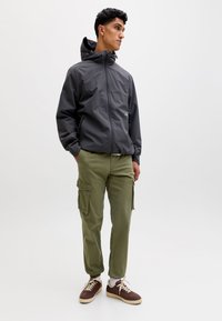 Uomo in piedi con le mani in tasca, indossa una giacca scura con cappuccio, pantaloni cargo oliva, calzini bianchi e scarpe da ginnastica marroni.