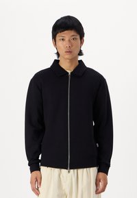 Veste zippée noire avec un col, fabriquée dans un matériau doux. Elle présente des manches longues et une coupe ajustée. Portée par-dessus une chemise de couleur claire.