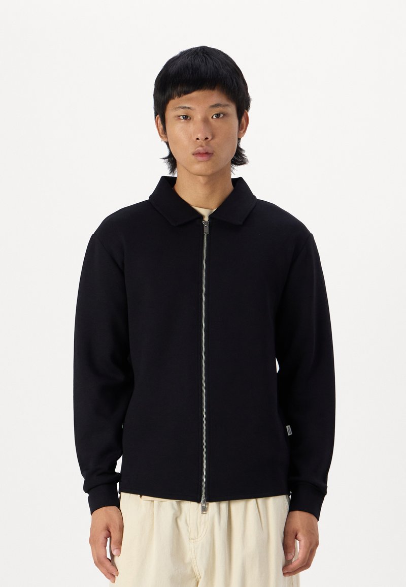 Veste zippée noire avec un col, fabriquée dans un matériau doux. Elle présente des manches longues et une coupe ajustée. Portée par-dessus une chemise de couleur claire.