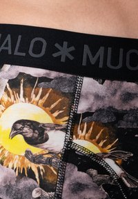 Fekete, rugalmas derékpánt "MUCHO" felirattal. Az anyagot színes madárgrafika díszíti, nap és felhők mellett. A textúrázott varrás fokozza a dizájnt.