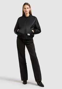 Schwarze Bomberjacke mit durchgehendem Reißverschluss, seitlichen Taschen, gerippten Bündchen und Kragen. Kombiniert mit hochgeschnittenen schwarzen Jeans und Stiefeln mit Absatz.
