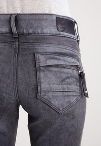 Jeans de mezclilla gris con una textura suave, bolsillo trasero con cremallera y parche de marca en la cintura, con un diseño ajustado.