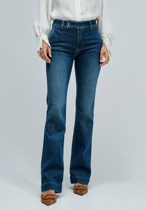 Mujer lleva jeans oscuros de talle alto con un ligero acampanado, combinados con una blusa blanca y zapatos de tacón en punta marrones.