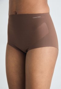 Chiloare maro Calvin Klein cu talie înaltă, cu o textură netedă și un logo subtil pe brâu; concepute pentru confort și fit.