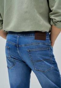 Vaqueros de mezclilla azul con un corte relajado, presentan bolsillos traseros y una etiqueta de cuero marrón con "Salsa Jeans". Textura suave y costuras detalladas.