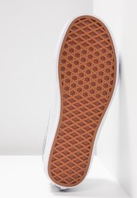 Semelle en caoutchouc avec un motif géométrique texturé ; dessus blanc avec des accents métalliques ; vue en angle montrant le détail et la forme de la semelle.