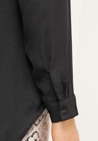 Camicia nera in satin a maniche lunghe con polsini abbottonati, caratterizzata da una texture liscia e una leggera lucentezza, accanto a un tessuto con stampa pitonata.