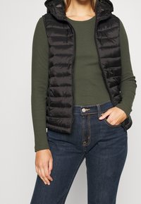 Svart vadderad väst med huva, som har dragkedja, bärs över en mörkgrön långärmad topp och blå jeans med kontrasterande stygn.