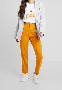 Grå rutigt blazer, vit grafisk t-shirt och knallorange byxor med en figurnära design. Avslutad med vita sneakers.