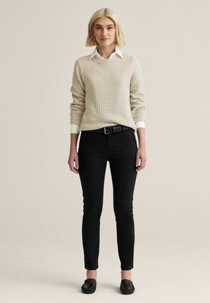 Nuori nainen, jolla on vaaleat hiukset ja beige teksturoitu neule, valkoinen kauluksellinen paita, mustat housut, musta vyö ja mustat loafers-kenkä, seisoo.