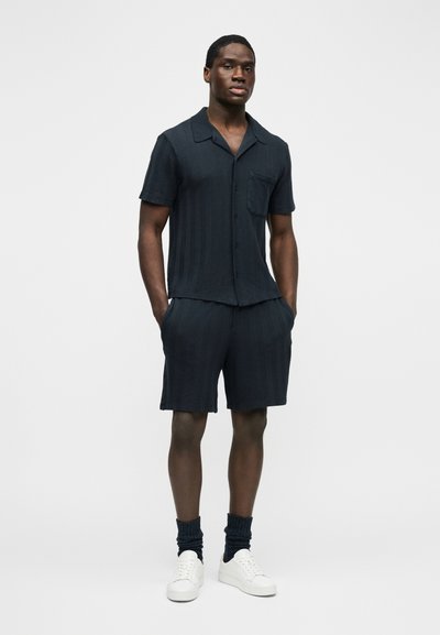 Homme debout les mains dans les poches, portant une chemise à manches courtes texturée sombre avec des boutons, un short assorti, des chaussettes foncées et des baskets blanches sur fond blanc.