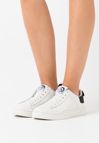 Białe sneakersy z gładkiej skórzanej cholewki, elastycznymi panelami po bokach, czarnym akcentem na pięcie i logo na języku. Płaska gumowa podeszwa.