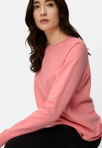 Hellrosa Sweatshirt mit langen Raglanärmeln und Rundhalsausschnitt, mit strukturierter Oberfläche und kleinem Logo nahe der Brust.