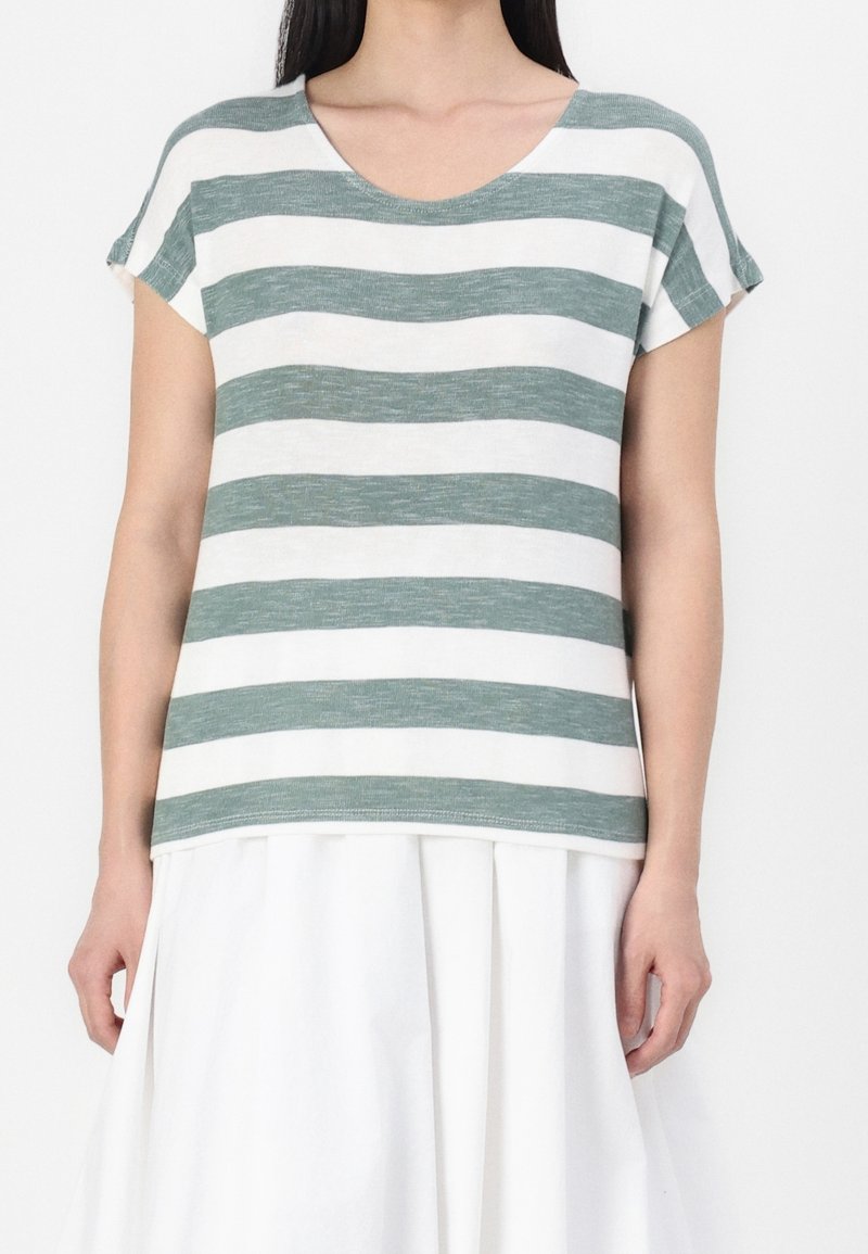 Femme portant un haut à manches courtes rayé horizontalement vert et blanc et une jupe blanche, debout devant un fond uni.