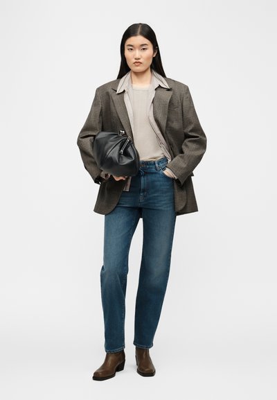 Γυναίκα όρθια, φοράει γκρι υπερμεγέθη σακάκι, μπεζ πουκάμισο, μπλε τζιν, καφέ μποτάκια αστραγάλου, κρατώντας μαύρη δερμάτινη τσάντα χειρός στο δεξί χέρι.