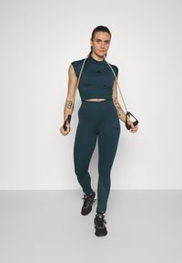 Top de sport scurt de culoare teal și colanți cu talie înaltă cu un design fără cusături, asociați cu adidași negri și benzi de rezistență în mână.