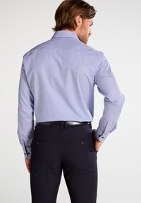 Camicia a righe blu e bianche con maniche lunghe, polsini con bottoni e colletto classico; indossata con pantaloni scuri; tessuto dalla consistenza liscia.