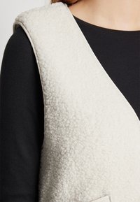 Krämfärgad väst i en mjuk, fleece-liknande material. Har en kontrasterande svart skjorta under med ribbad textur.