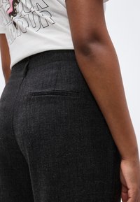 Κοντινό πλάνο ενός ατόμου που φορά σκούρα γκρι παντελόνια με υφή και πίσω τσέπη, καθώς και λευκό γραφικό T-shirt.