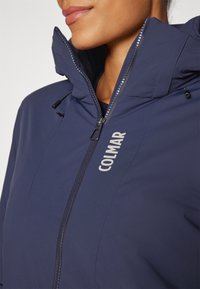 Navy blue waterproof jacket with a high collar, zipper closure, and elastic drawstrings. Features white "COLMAR" logo on chest.