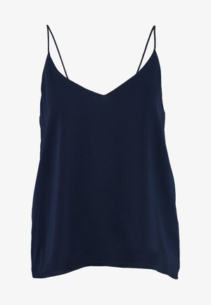 Femme portant un haut sans manches bleu marine, un jean noir retroussé, des chaussures grises à motif serpent et un sac à dos en cuir noir sur l'épaule.
