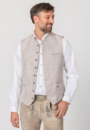 Mann trägt ein weißes Hemd, eine hellgraue gemusterte Weste und traditionelle beige bestickte Lederhosen, schaut nach rechts und lächelt.