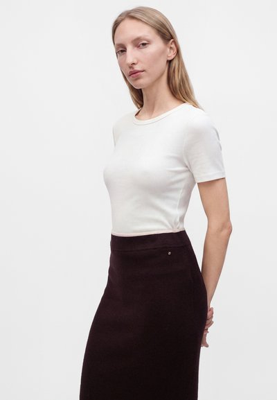 Jeune femme aux cheveux blonds raides portant un haut blanc à manches courtes et une jupe sombre taille haute, debout devant un fond uni.