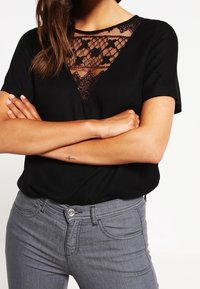 Top negro de manga corta con un inserto de encaje en forma de V en el escote. Combinado con jeans grises que tienen bolsillos frontales y cierre de botón.