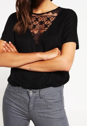 Sort kortærmet top med et V-formet blondeindsats ved halsudskæringen. Sammen med grå jeans med forlommer og knaplukning.