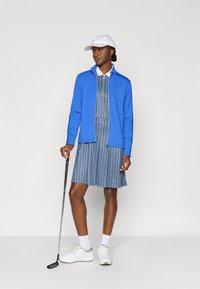 Zila sporta jaka pāri rakstainai zilai kleitai, balta cepure un balti sporta apavi. Turot golfa nūju, stāvot pret gaišu fonu.