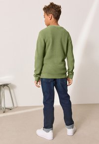 Vihreä neulepaita, väljällä istuvuudella, pitkät hihat ja resorit manseteissa, yhdistetty tummansinisiin farkkuihin ja valkoisiin tennareihin. Yksinkertainen, teksturoitu muotoilu.