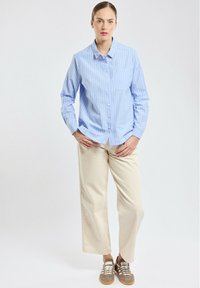 Chemise boutonnée rayée bleu clair avec une poche, associée à un pantalon large beige et des baskets décontractées marron. Arrière-plan neutre.