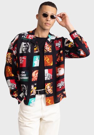 Czarna koszula z guzikami, ozdobiona kolorowymi nadrukami w stylu pop art przedstawiającymi twarze i tekst, z długimi rękawami i luźnym krojem, w połączeniu z kremowymi spodniami.