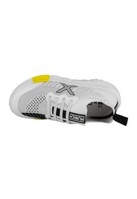 Zapatilla deportiva de malla blanca con acentos en negro, con una prominente puntera amarilla, cierre de cordones y un tirador en el talón.