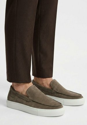 Zapatillas de hombre de ante marrón sin cordones con suelas de goma blancas, usadas con pantalones a medida de color marrón oscuro.