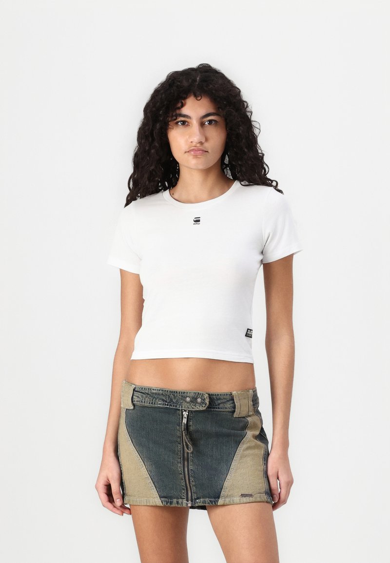 T-shirt bianca corta con maniche corte e un piccolo logo nero. Abbinata a una minigonna di jeans con pannelli colorati e dettagli con zip.