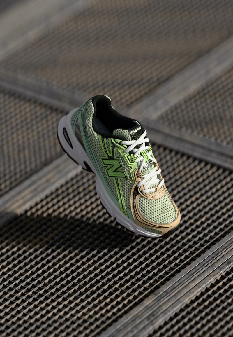 Chaussure de sport verte et beige avec des lacets blancs et le logo "N" sur un fond de grille métallique texturée.