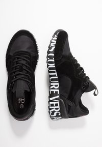 Svarta snörskor med tjocka vita sulor som har fet text "VERSACE COUTURE", sidoband, och blandade mocka- och tygpaneler.