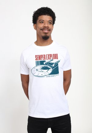 Man draagt een wit T-shirt met een ruimteschipgrafiek en rode tekst "SEMPER EXPLORO" en staat tegen een witte achtergrond.