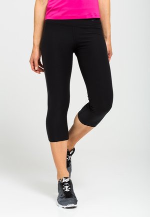 Kobieta ubrana w czarne skrócone legginsy, różowy sportowy top i czarno-białe buty do biegania, stojąca z jedną lekko uniesioną stopą.