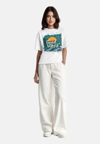 Witte t-shirt met een kleurrafische graphic van golven en een zon, tekst luidt "Good Vibes!" Model draagt losse witte broek en sneakers.