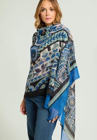 Châle bleu et noir à motifs floraux et géométriques, tissu léger, design drapé, bords à franges, porté par-dessus un jean en denim.