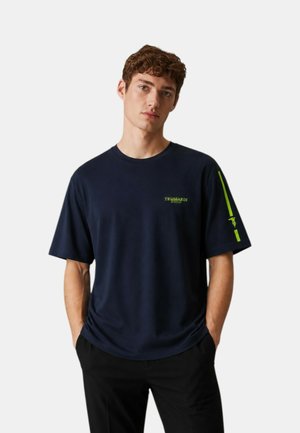 Giovane uomo con capelli ricci che indossa una maglietta blu navy con logo Trussardi Swim verde e pantaloni neri, mani in tasca, espressione neutra.