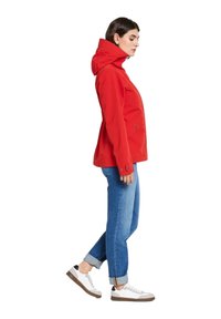 Veste imperméable rouge avec capuche, poches latérales et détails en bouton, associée à un jean bleu clair retroussé et à des baskets blanches.